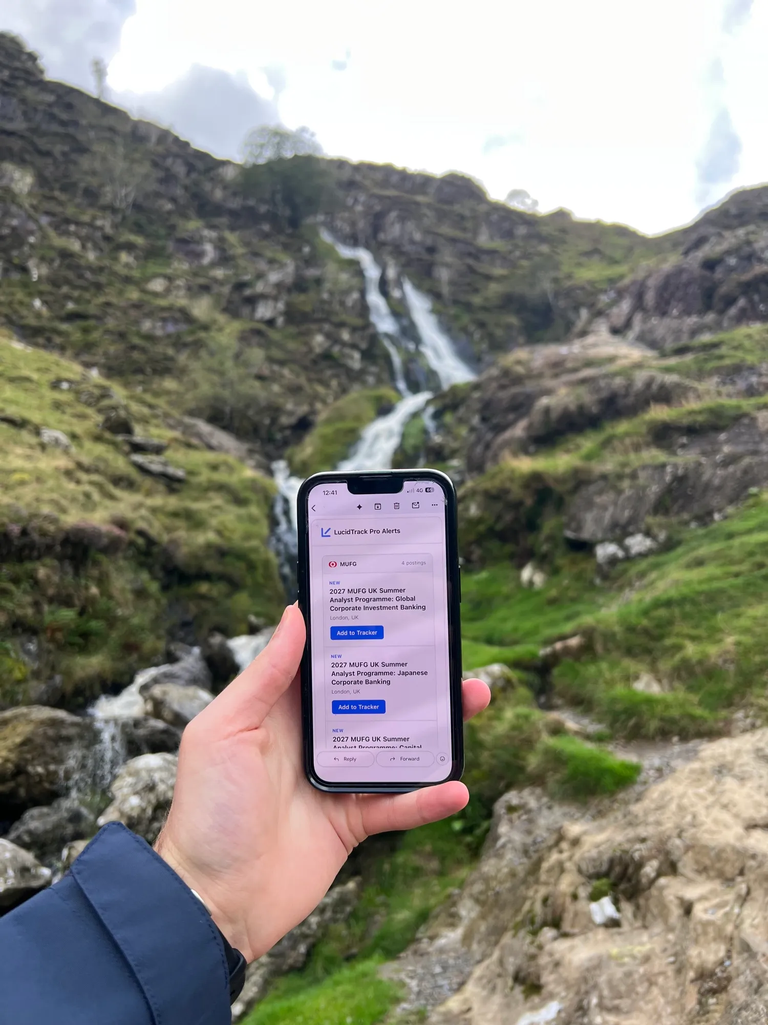 A phone held up in front of a waterfall in the Lake District, showing a LucidTrack Pro Alerts email with new roles to add to the tracker.