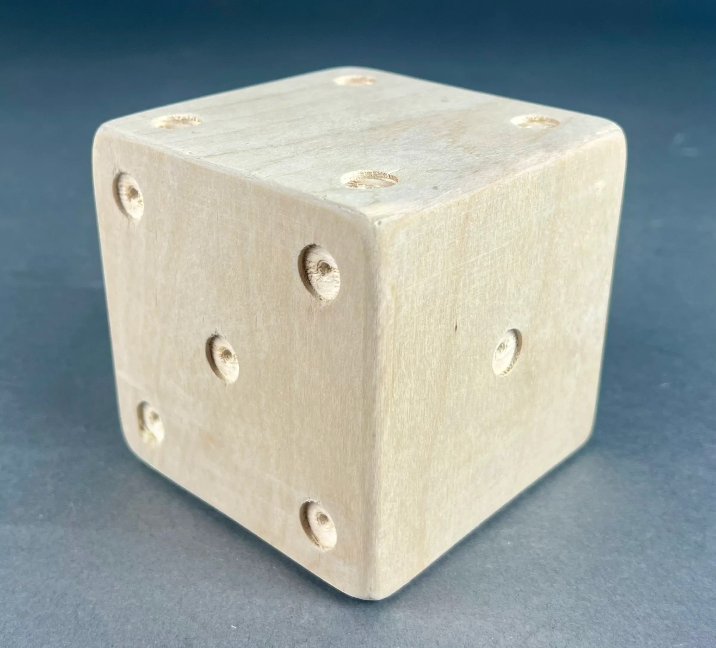 The finished die: a pale plywood cube with drilled pips, on a blue-grey backdrop.