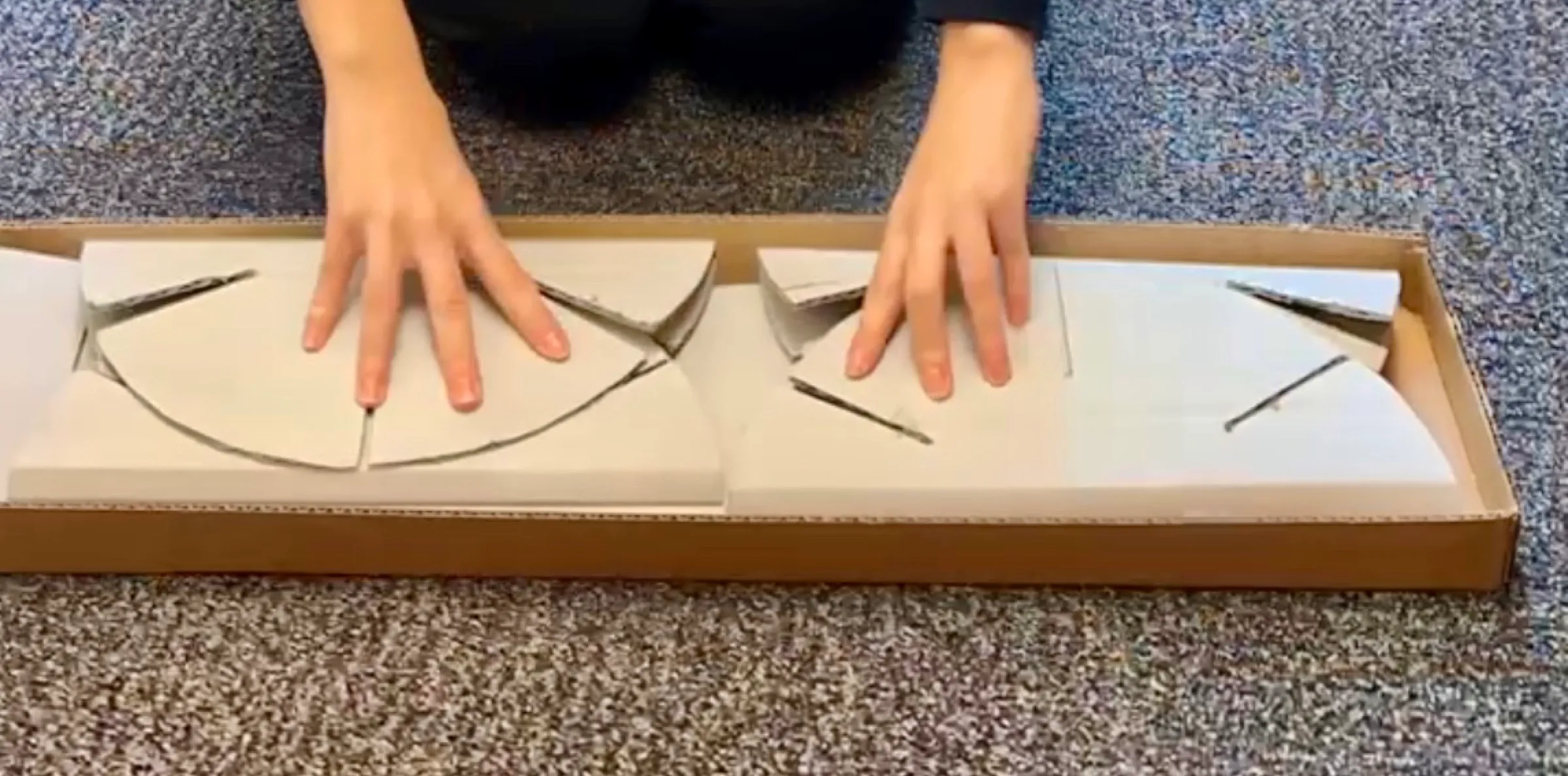 Hands pressing the flat-packed cardboard parts into a long, shallow box on a carpet.