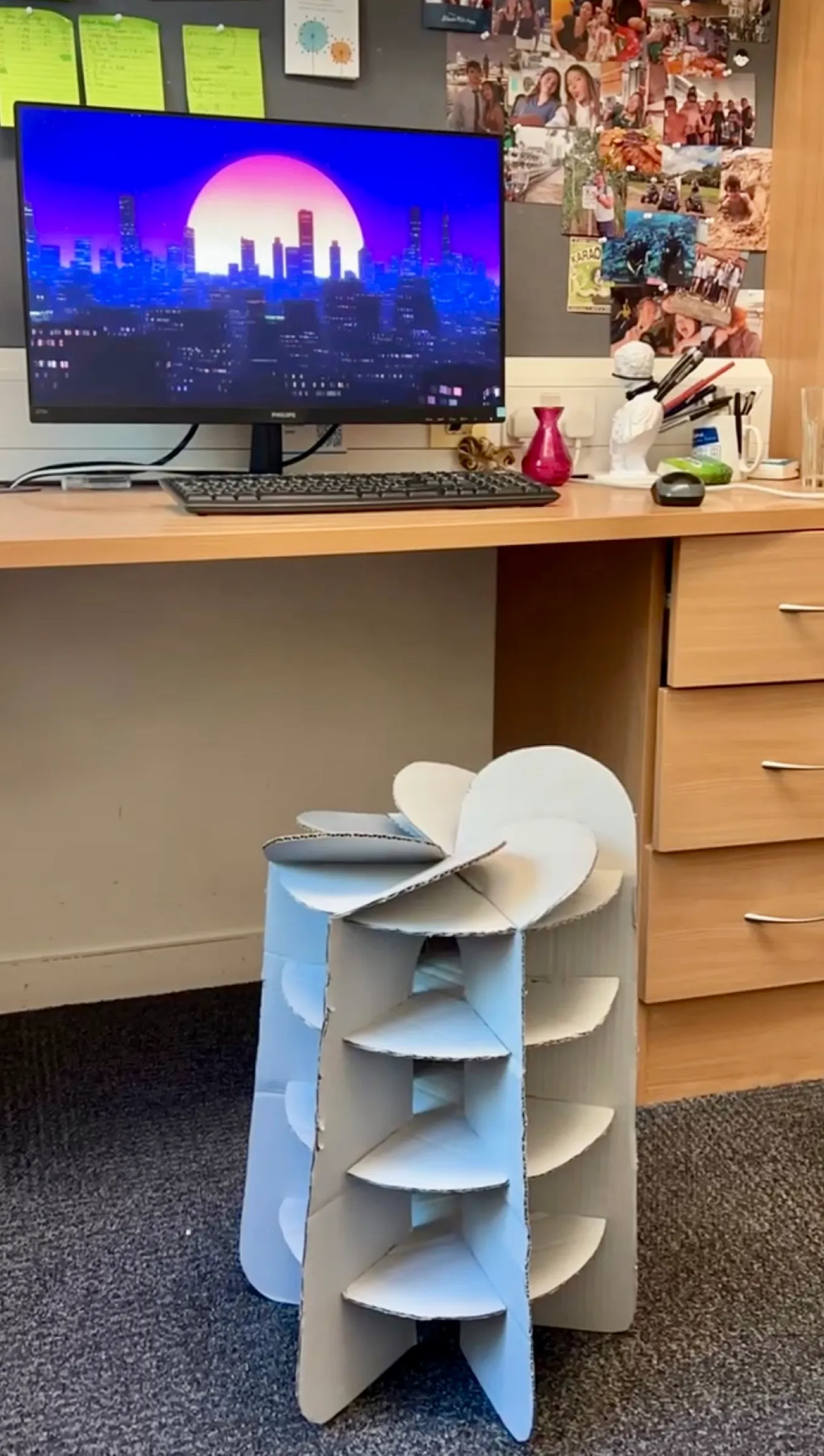 The grey stool beside an office desk with a monitor and a pinboard of photos.