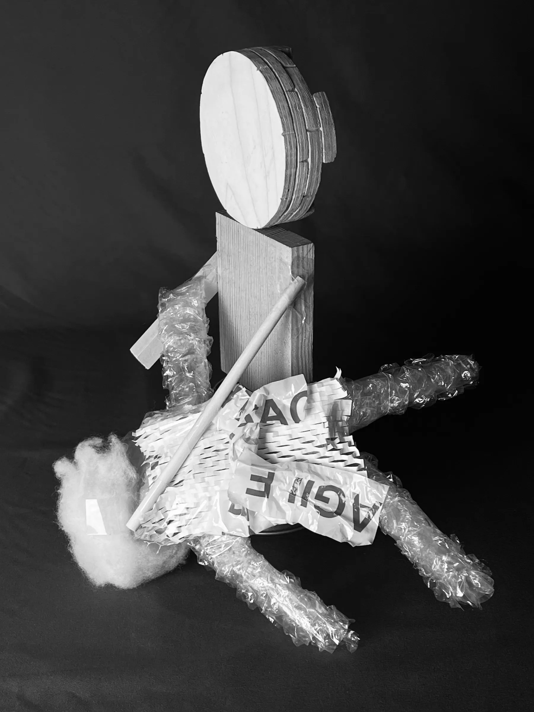 Black and white photo of a model figure: a round wooden head on a block body, limbs wrapped in bubble wrap and packing paper, a dowel through the chest.