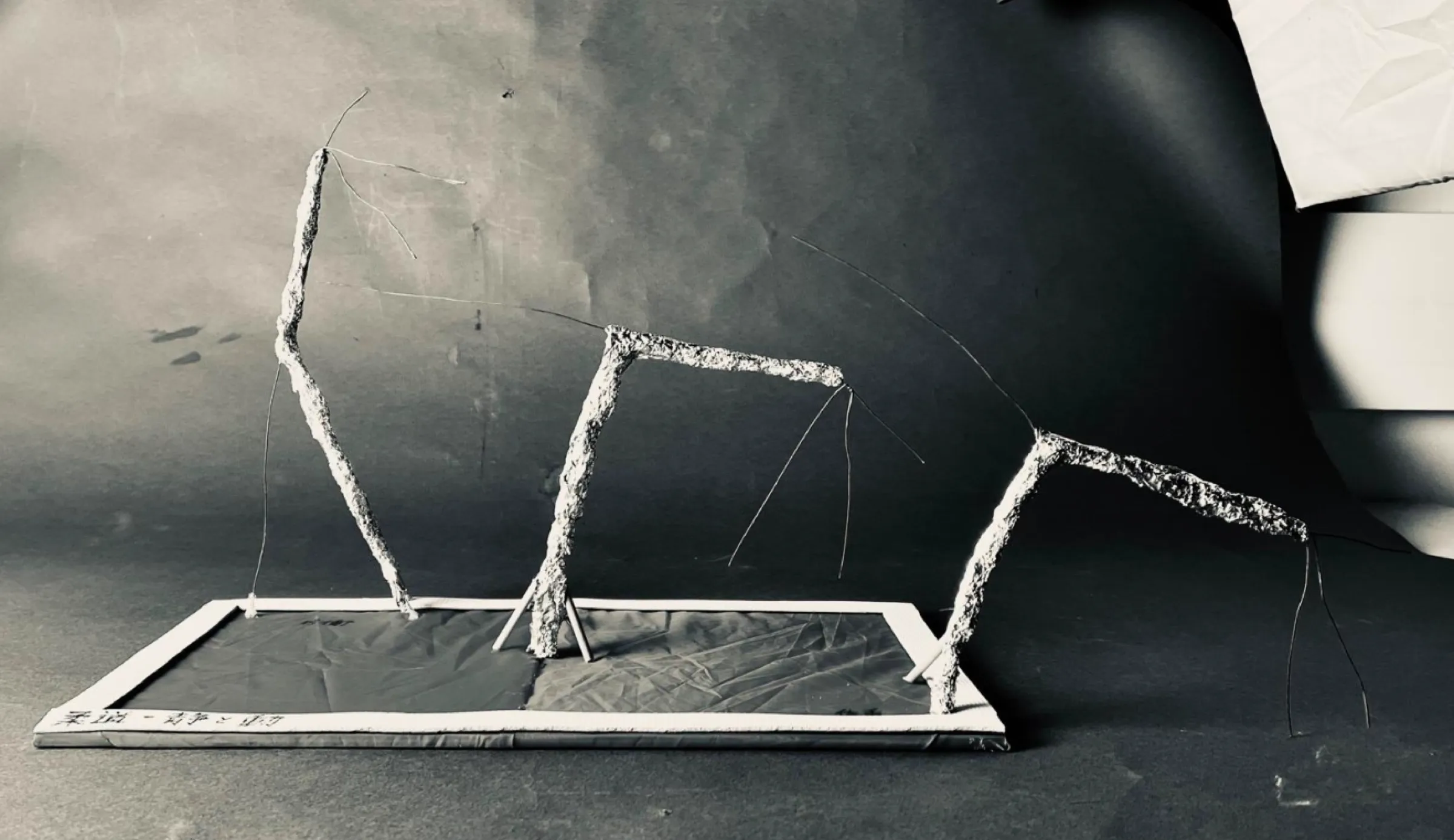 Black and white photo of a wire model on a flat base: three bent, plastered wire figures in a row, each leaning further over.