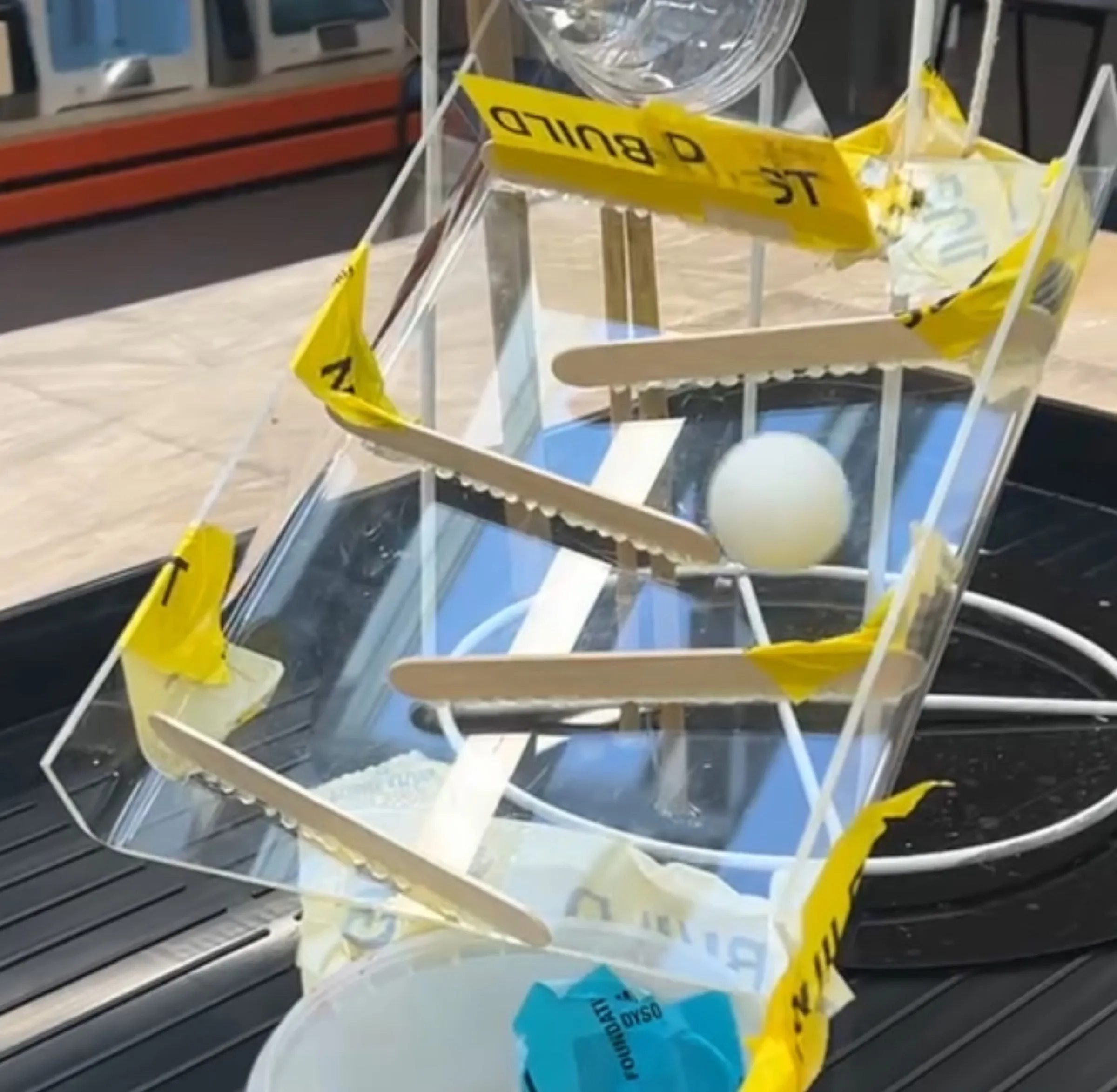Close-up of a clear acrylic ramp with wooden deflectors and yellow tape, a ping-pong ball rolling down it.