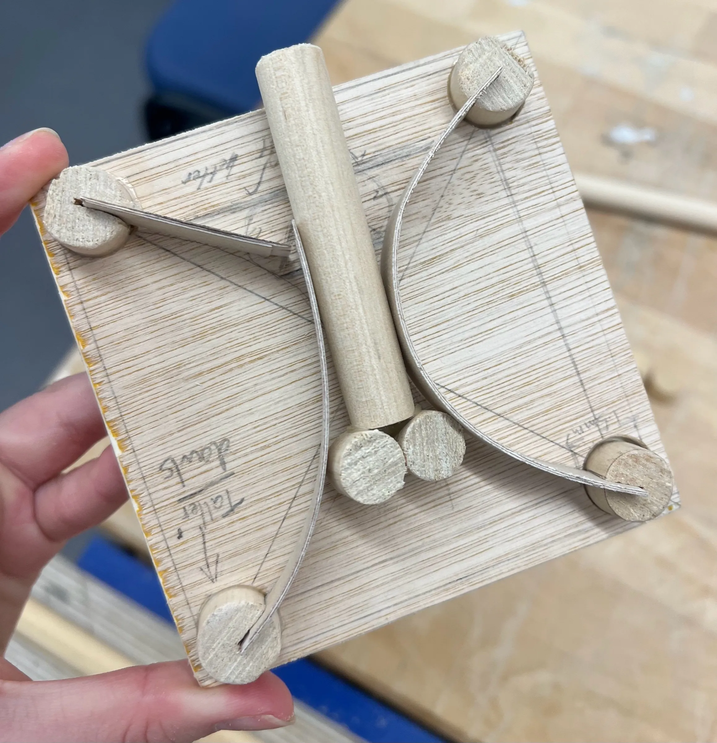 A plywood test piece held in a hand: a dowel post down the middle and two curved strips of thin wood running between corner dowels.
